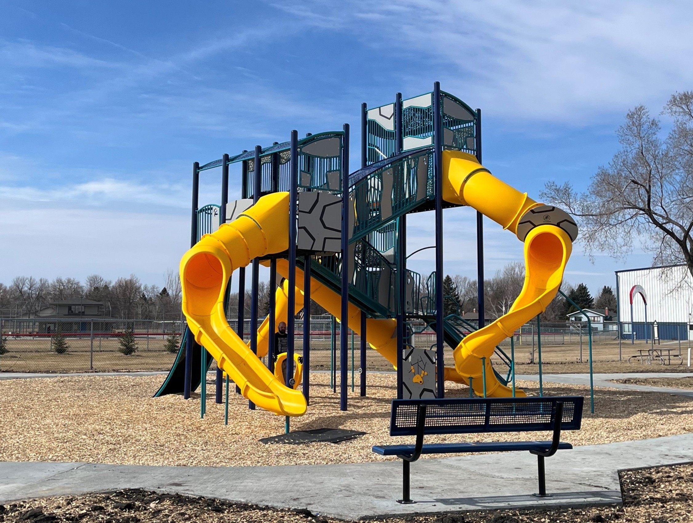 Navy and grey multi-level playground with four yellow tube slides and navy metal bench. 